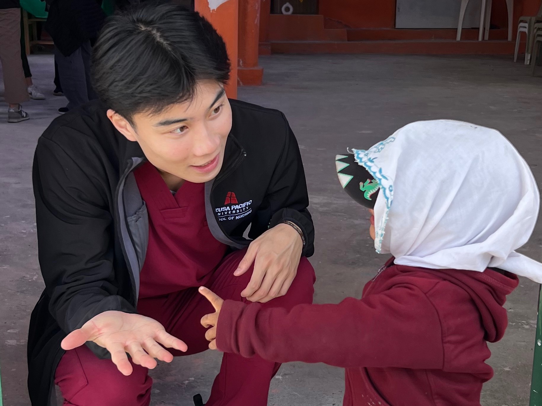 Wearing his nursing scrubs, Joshua Tran kneels down to talk to a child.