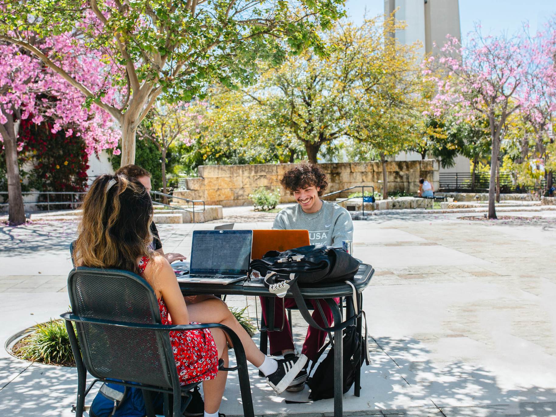Two students sit outside at a table with their laptops.