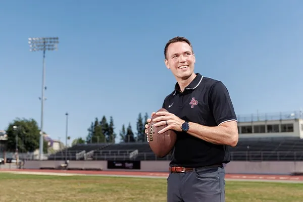 apu's new head coach holding a football and smiling