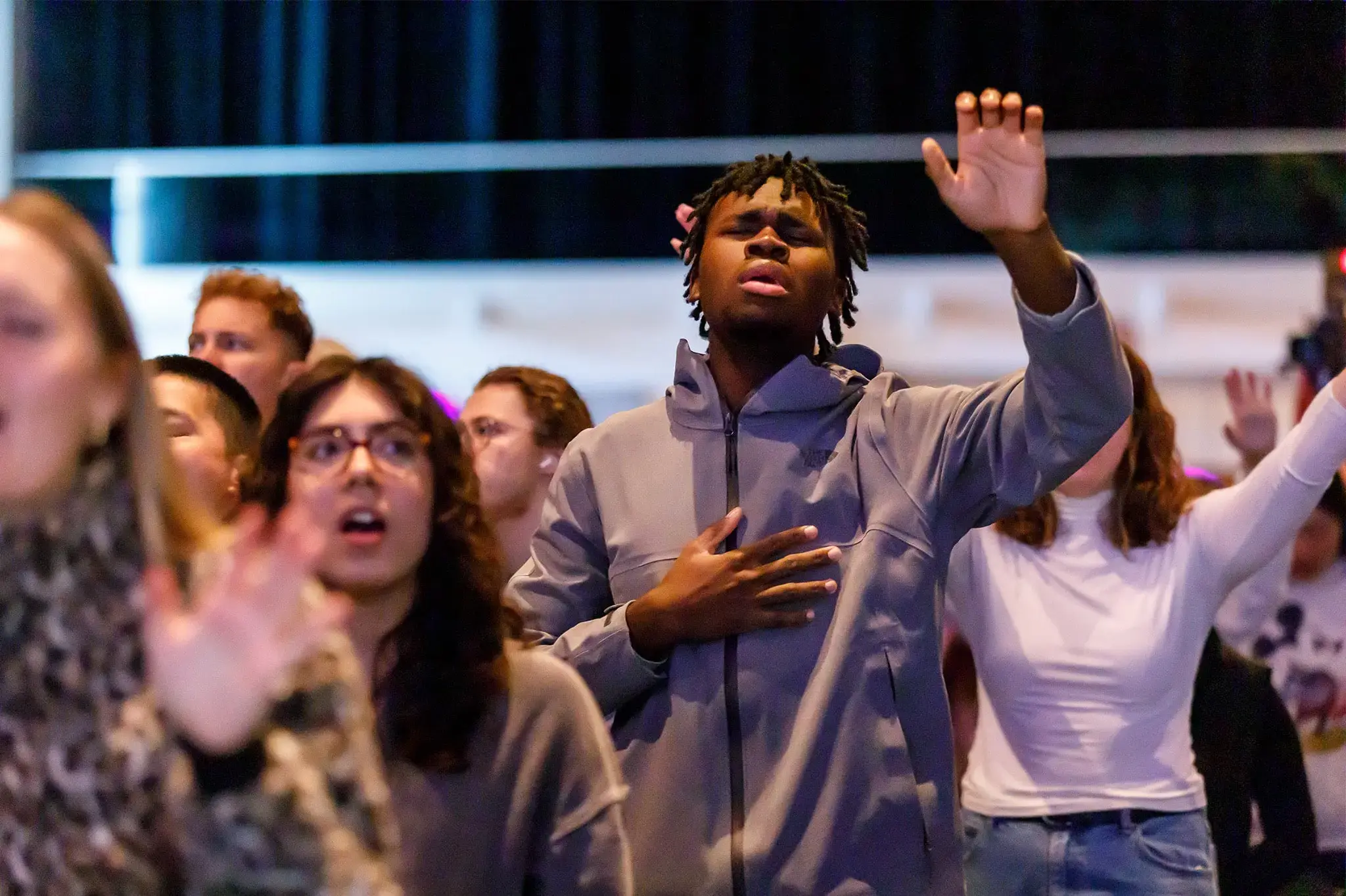 A young man in a gray hoodie with his hand on his heart and one hand raised, eyes closed, in a crowd.