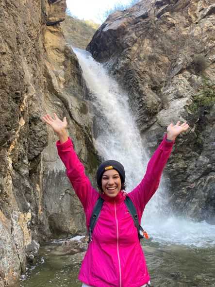 Ashley Jones standing with arms raised in joy and victory in front of a waterfall after a hike.