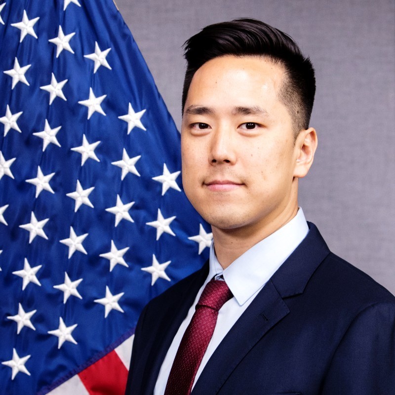 Daniel Lee government photo sitting in three-quarter profile in front of am American flag.