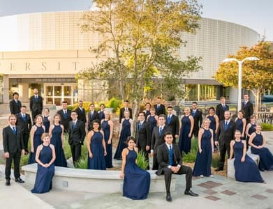 Image of the chamber singers in front of the darling library at APU wearing black suits and blue dresses.